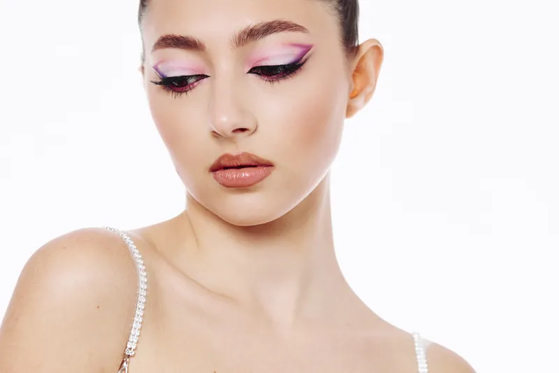 Young woman with artistic purple and pink eye makeup looks downward showing flawless skin and soft lips. Studio portrait with focus on beauty, fashion, and creative cosmetics.