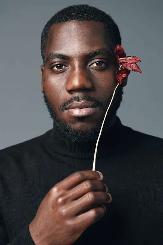 African man with dark skin holding dried flower near face in creative conceptual visual style. Portrait with serious expression and artistic mood on neutral background for artistic project.