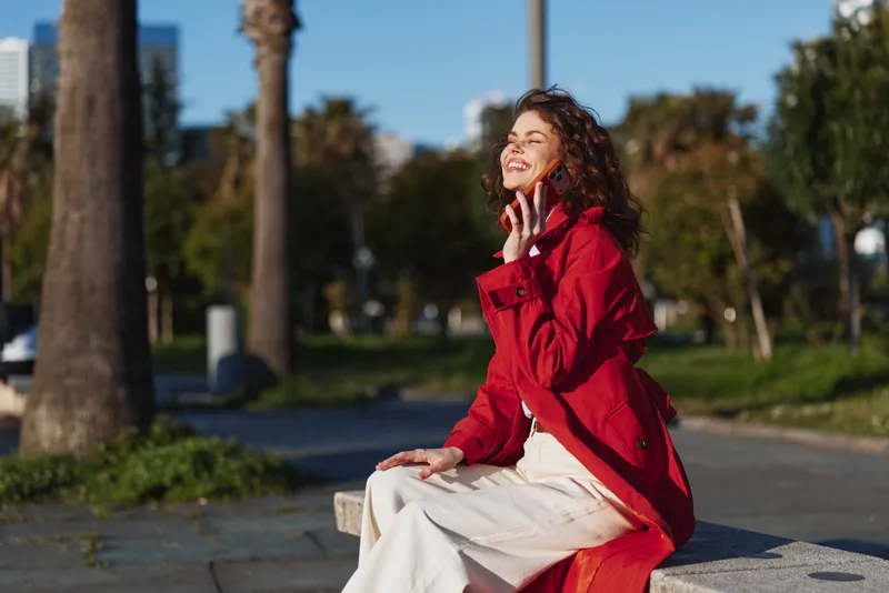 Young woman with curly hair wearing a red jacket enjoys a sunny day sitting on a bench in a park with a joyful smile and closed eyes.