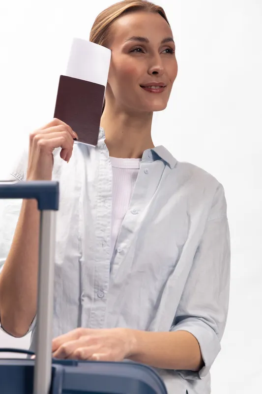 Young woman holding passport and ticket stands near suitcase on white background. Travel concept with casual clothing and positive expression preparing for trip or vacation.