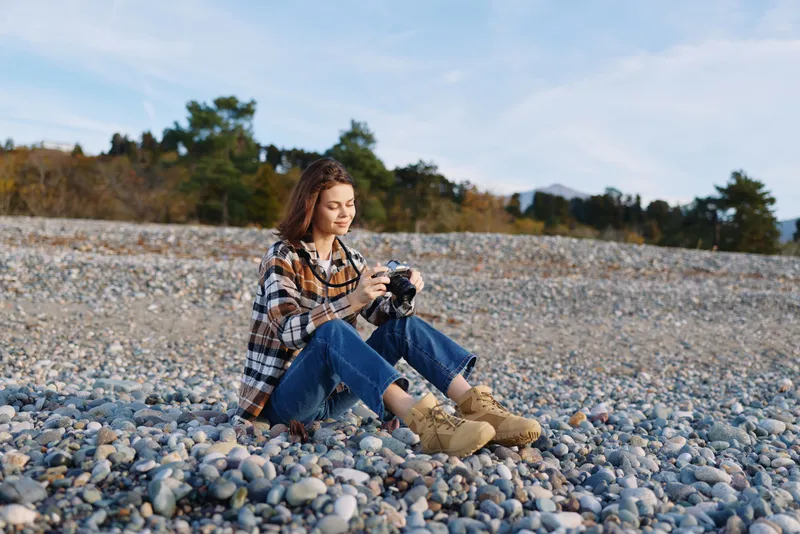 Woman photographer sits on a pebble beach, focused on her camera, in a casual plaid shirt and jeans, enjoying outdoor photography amid calm scenery and boots, a peaceful moment captured.