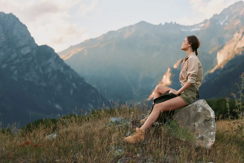 A woman in casual clothes sits on a rock in the mountains, enjoying the peaceful nature and fresh air during a sunny day. Concept for lifestyle and outdoor relaxation.