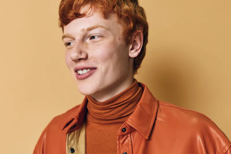Young man with red hair smiling and looking sideways wearing a brown turtleneck and orange leather jacket against beige background. Portrait of casual fashion style.