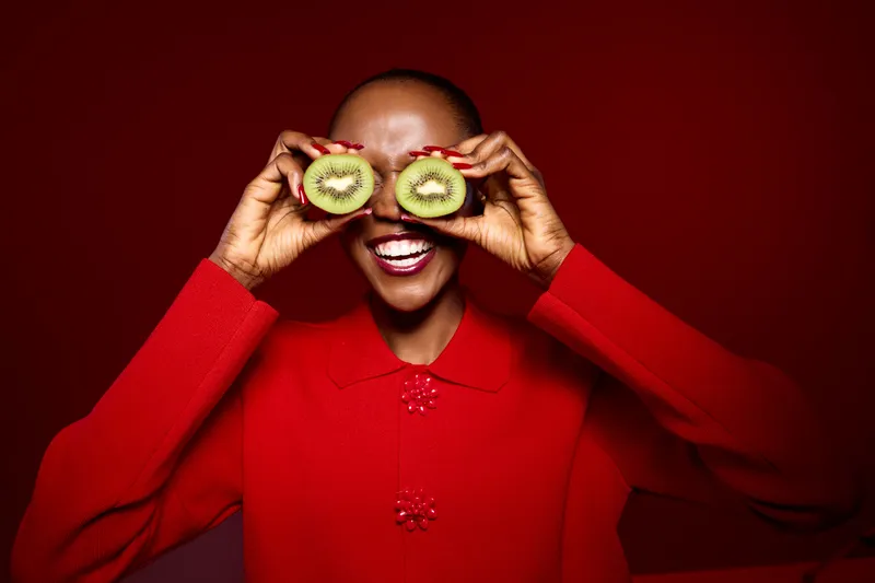 Smiling black woman with dark skin wears red sweater and holds two halves of kiwi fruit over eyes, posing against red background. Concept of healthy lifestyle, fruit, and joyful mood.