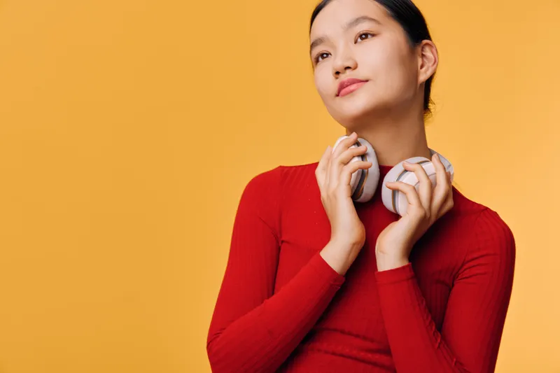 Woman in red sweater holding wireless headphones with hands near chin, posing with eyes looking up, thoughtful expression on yellow background in studio portrait.
