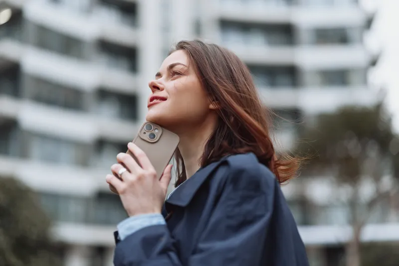 Young woman with long brunette hair holds a smartphone near her face while standing outdoors in a casual jacket with a modern building background.