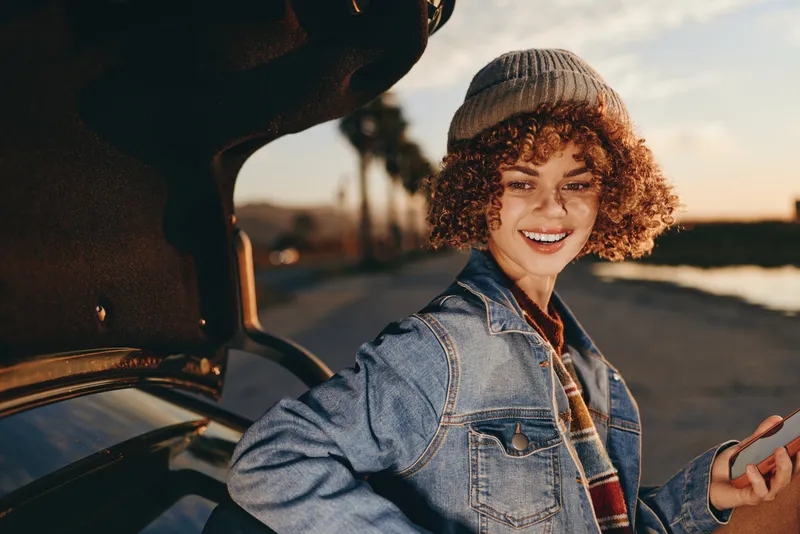 Woman lifestyle, denim jacket, curly hair, smiling near car window at golden sunset, relaxed posture, road outdoors, casual fashion, warm light, friendly expression, travel mood