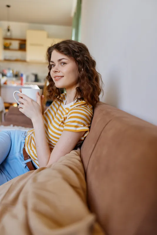 Cozy scene of a woman relaxing on a sofa at home, holding a mug and smiling softly as she enjoys a quiet morning in a sunlit living room with warm light and comfortable surroundings