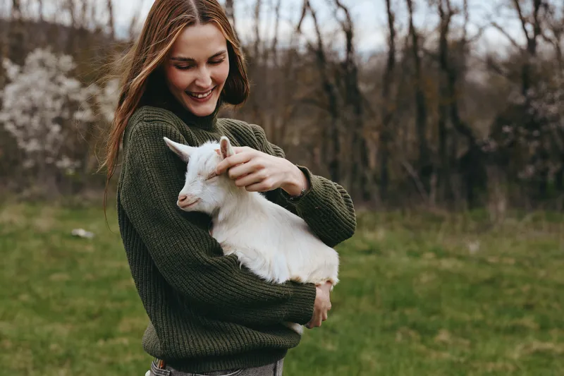 A young woman in a green sweater gently holds a white baby goat outdoors in a grassy field during early spring, smiling warmly while connecting with the animal.