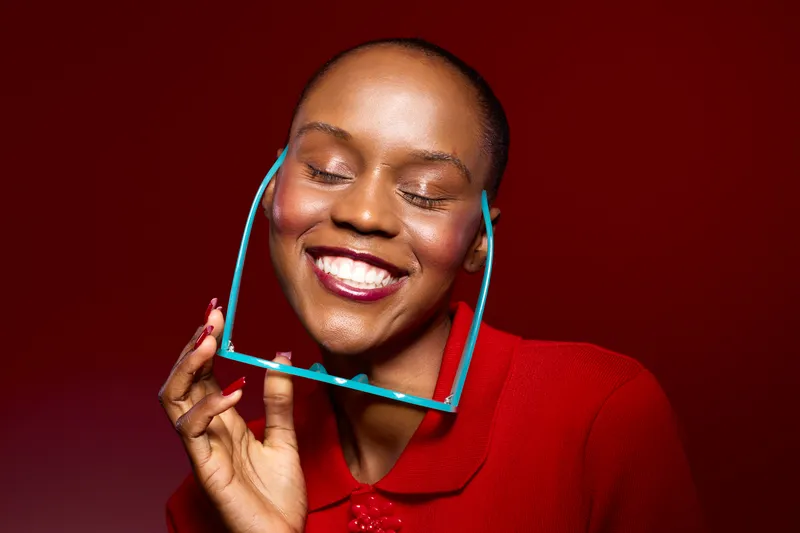 Smiling African woman with dark skin holding blue glasses near face, eyes closed, wearing red top. Studio portrait on red background. Concept for happiness, beauty, and fashion accessories.
