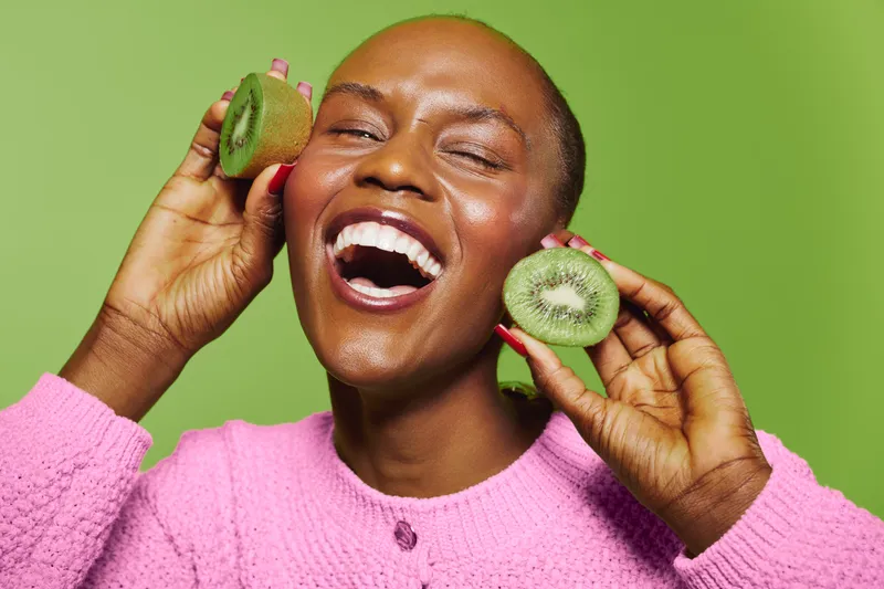 Happy black woman with dark skin holds fresh kiwi fruit near face, smiling with closed eyes on green background. Concept of healthy lifestyle, natural food, and joyful mood.
