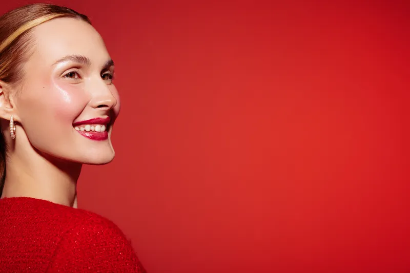Smiling woman with red lipstick and shiny skin looks sideways on red background. Concept of beauty, makeup, skincare, happiness, and confident female portrait in studio lighting.
