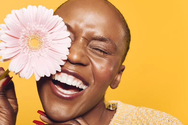 Joyful black woman with dark skin holds pink flower near face smiling with closed eyes on yellow background. Concept of happiness, beauty, skincare, and natural expression.
