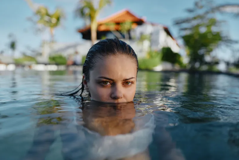 Young woman partially submerged in clear water at a tropical resort, smiling with a relaxed expression as the sun warms her face and palm trees hover in the background