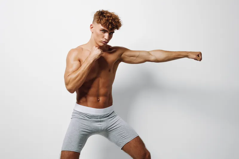 Young athletic man with curly hair practicing boxing punch in gray shorts on white background. Muscular body, fitness training and workout for strength and health concept.