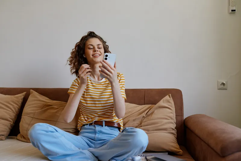 A smiling woman sits on a cozy sofa at home, scrolling on a smartphone. Casual striped tee and jeans create a relaxed, everyday mood for woman lifestyle and home concepts.