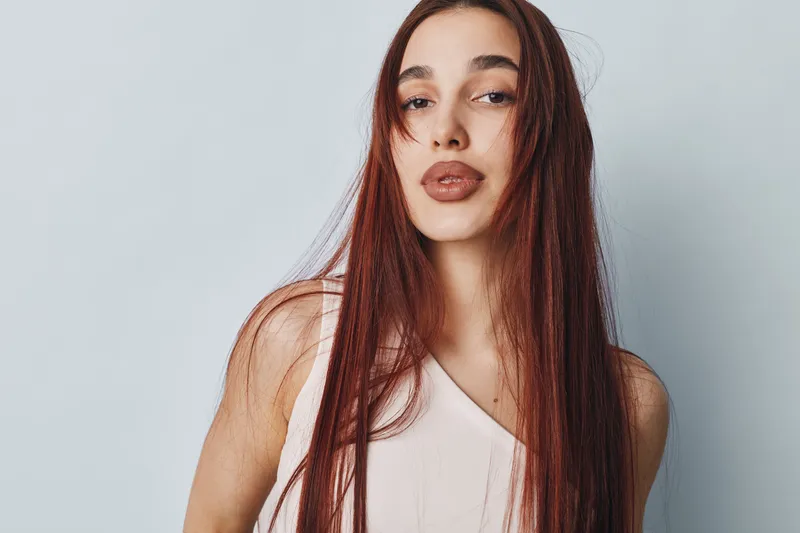 Young woman with long red hair and natural makeup posing against plain light background wearing sleeveless top. Portrait of confident female with neutral expression and loose hair.