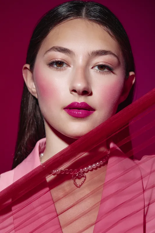 Close-up of a young woman with bold pink lipstick and elegant pearl necklace posing behind sheer pink fabric. Studio portrait on magenta background. Concept of beauty, fashion, and creative