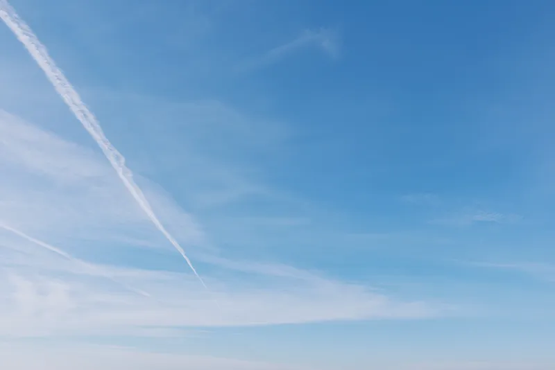Blue sky with a single white contrail slicing diagonally, wispy clouds near the horizon, bright daytime light, clean open air, and a tranquil expansive atmosphere for travel, aviation, and nature