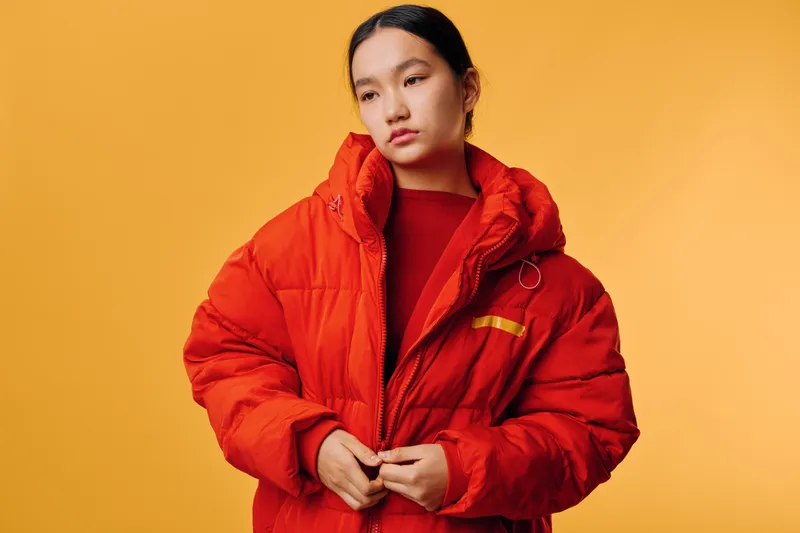 Young woman wearing bright red puffer jacket standing against orange background looking away with neutral expression and hands adjusting zipper for warm winter fashion style.