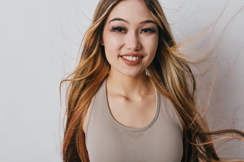 Young woman with long hair and smile wearing sleeveless top on gray background. Portrait of happy female with natural makeup and casual hairstyle in studio.