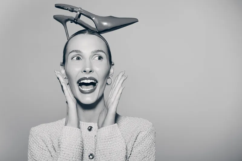 White woman with fair skin and red hair laughs holding high heel shoe on head in studio. Concept of fashion humor and playful portrait.