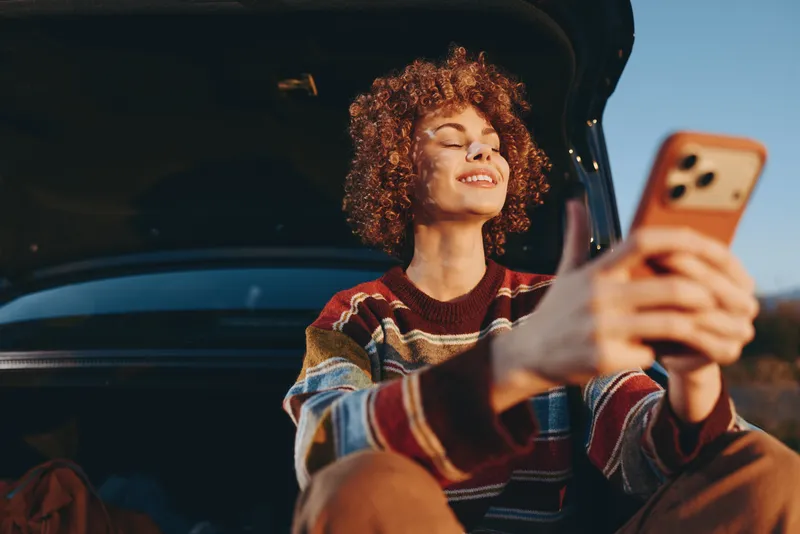 Woman selfie smartphone car lifestyle, while sitting in car at golden hour, curly hair smiling, casual sweater, relaxed lifestyle moment, warm sunset light, outdoors background, travel vibe
