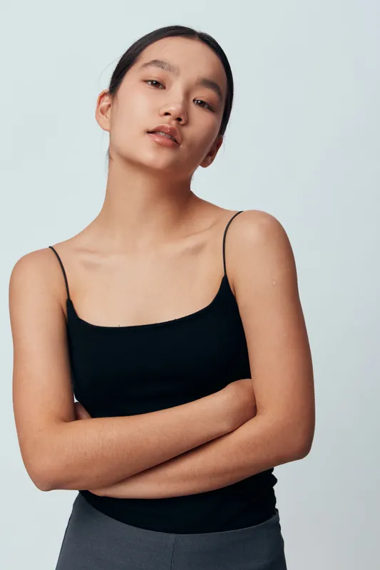 Young woman with arms crossed wearing black sleeveless top looking confidently at camera on light background. Casual fashion portrait with natural makeup and minimalistic style.
