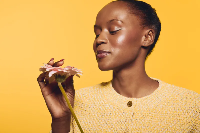 Portrait of a young black African woman with dark skin holding a pink flower, wearing a yellow sweater on a yellow background. Concept of natural beauty and floral wellness.