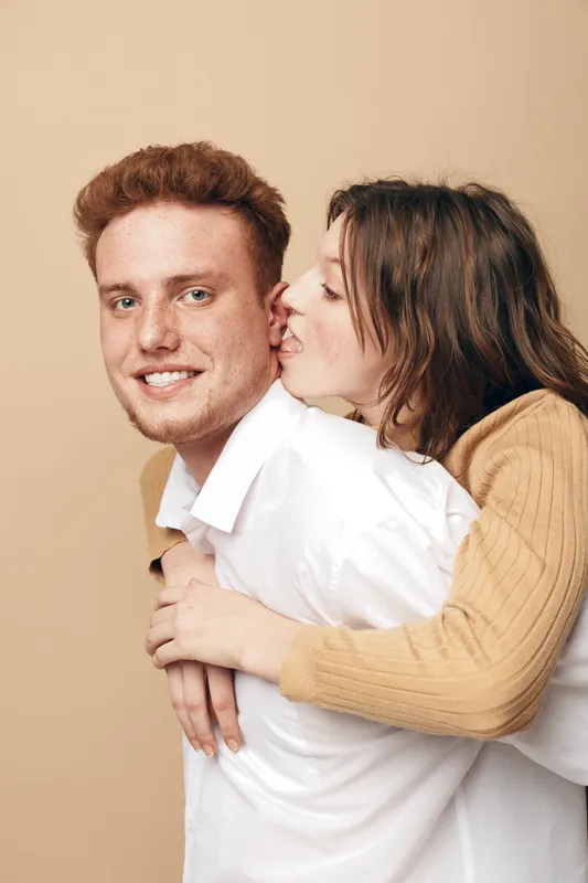Happy young couple in casual clothes share affectionate moment, woman playfully kisses man on cheek. Warm beige background creates cozy and intimate atmosphere of love and friendship.