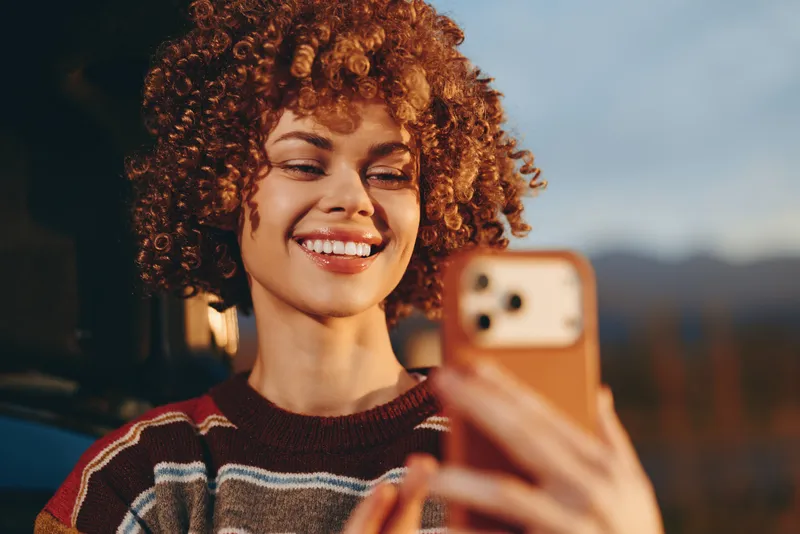 Woman lifestyle selfie with smartphone, curly red hair, smiling brightly while holding phone outdoors during golden hour, close portrait with soft blurred background, friendly casual mood