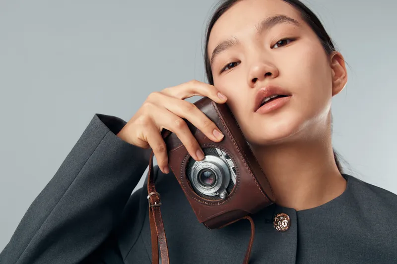 Woman holding vintage camera close to face with stylish gray jacket. Elegant portrait photography with minimal background for retro fashion and creative concept in studio.