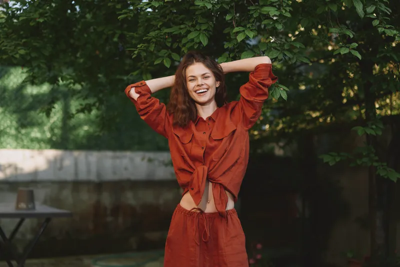 Young woman with curly brown hair laughs with arms raised in a rust colored matching set outdoors, enjoying a carefree moment in dappled shade, sunlight warming her bright smile on a summer day