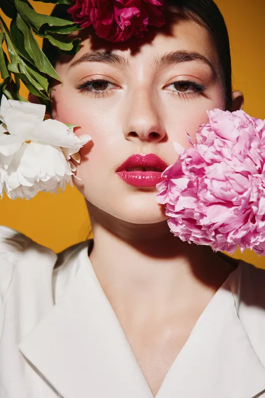 Young woman with glossy pink lips and soft makeup posing with white and pink peonies on her face. Fashion portrait on bright yellow background. Concept of beauty, floral, and creative style.