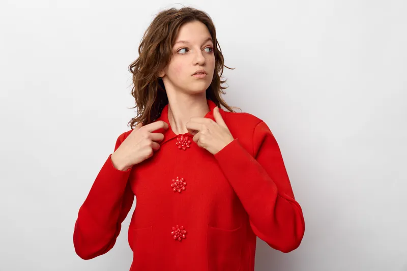 Young woman in red knitted sweater looks sideways with thoughtful expression. Casual fashion, autumn style, and natural beauty concept on plain white background in studio lighting.