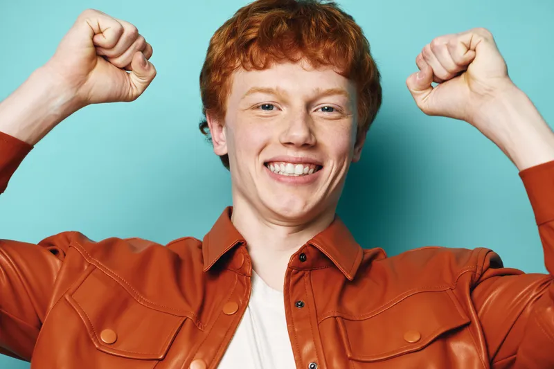 Young man with red hair smiling and raising fists in victory gesture wearing brown jacket against blue background. Portrait of confident happy male with positive emotion.