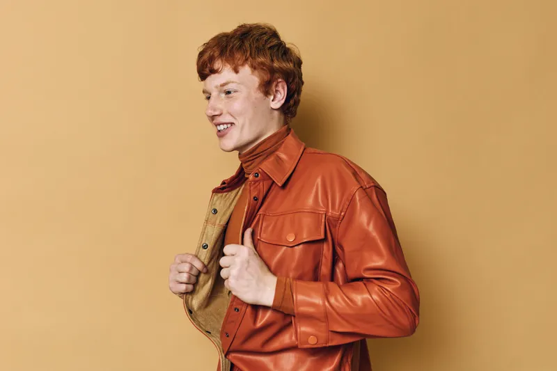 Young man with red hair wearing brown leather jacket and turtleneck smiling, standing in front of plain beige background for fashion and lifestyle portrait.
