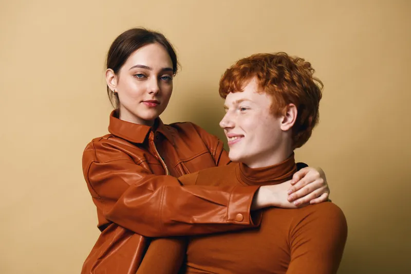 Young couple with brown hair and casual outfits embracing each other on beige background. Woman with leather jacket holding smiling redhead man in close pose.