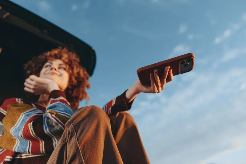 Woman with curly hair taking a selfie, holding a smartphone outdoors, wearing a cozy striped coat, looking up to blue sky, casual lifestyle, candid windy moment, bright natural light