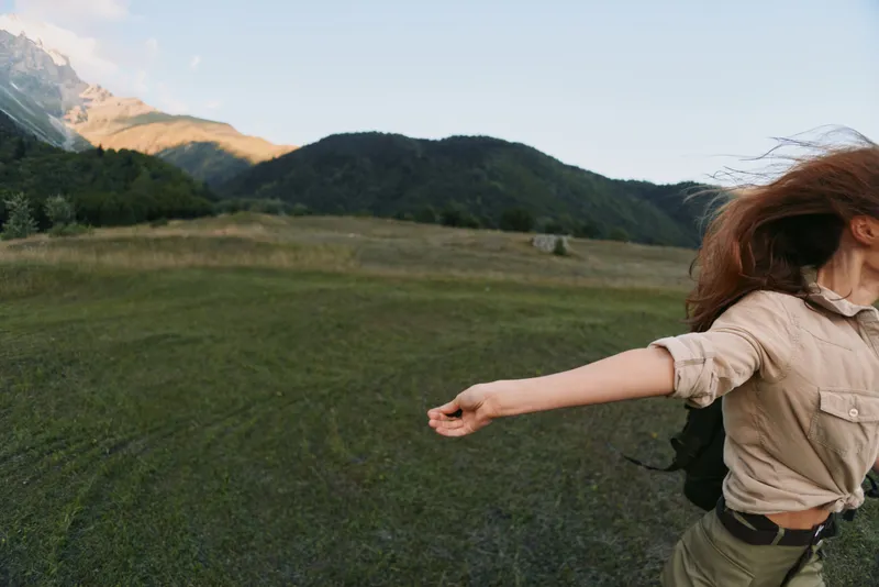 Woman in casual beige shirt enjoys expansive outdoors, arms spread, open field with mountain backdrop, conveying freedom and carefree lifestyle moment, under clear sky, warm light, moving wind, sense