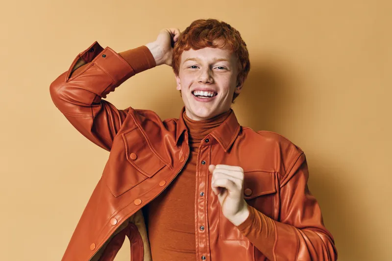 Young man with red hair smiling while wearing brown leather jacket and turtleneck shirt against beige background in studio portrait
