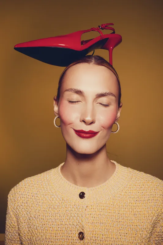 Woman with closed eyes and red lipstick balances red high heel shoe on head wearing yellow textured sweater and gold hoop earrings. Concept of fashion, style, and playful elegance.