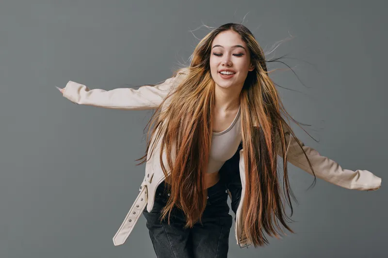 Young woman with long hair and casual jacket posing with arms outstretched against gray background smiling. Studio shot, fashion, movement, happy expression on face.