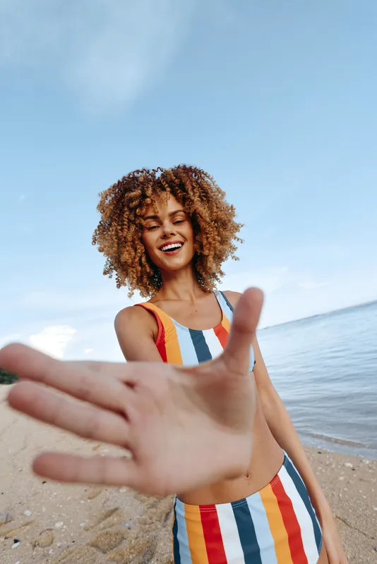 Joyful woman with curly hair stands on sandy beach by ocean, smiling at camera, raises one hand in foreground as greeting, colorful striped swimsuit, bright summer sky, travel vacation