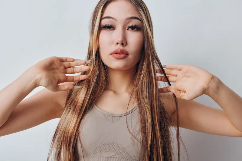 Woman with long straight hair posing and looking at camera with hands near head wearing sleeveless top on light background for beauty and fashion portrait.