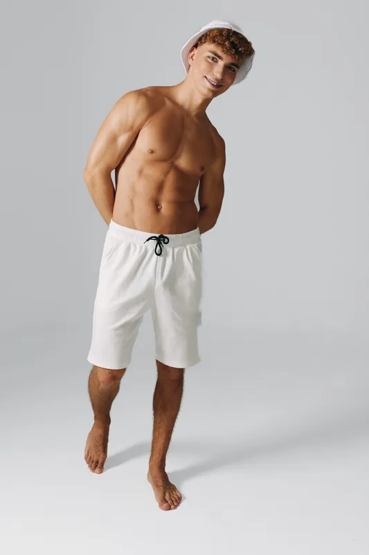 Young man with muscular body wearing white shorts and bucket hat standing barefoot on gray background with hands behind back and smiling at camera.