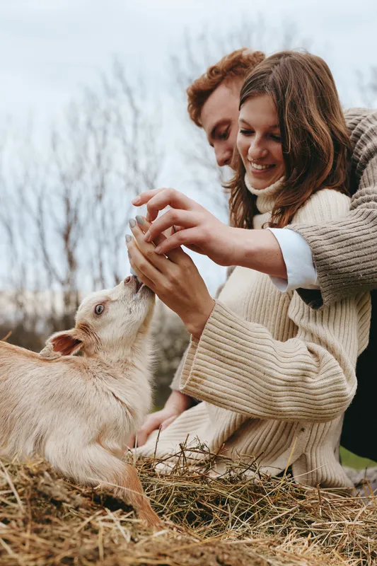 A joyful lifestyle moment of two people sharing a gentle interaction with a curious goat kid outdoors, capturing bonding, care, and playful affection in a natural farm setting.