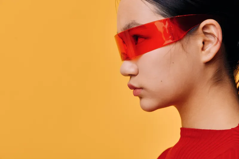 Young woman in red futuristic sunglasses and matching red top in closeup profile on orange background. Stylish female model with dark hair and minimalistic modern look in studio.
