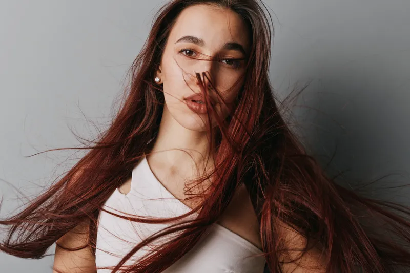 Portrait of young woman with long flowing hair and natural makeup posing against gray background. Female model looking at camera with confident expression and casual outfit.