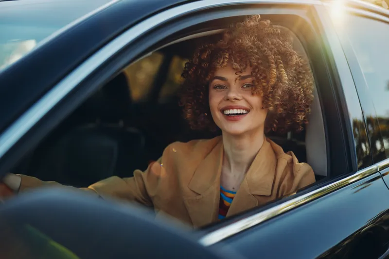 Woman in a retro car smiles warmly, enjoying a sunny drive, wearing a blazer, showcasing curly hair, candid lifestyle moment with vintage film color and soft glow